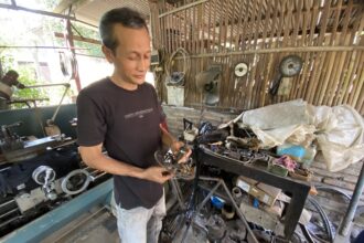 Man wearing a dark T-shirt stands in a cluttered workshop, holding bicycle parts over a workbench with tools and machinery nearby.