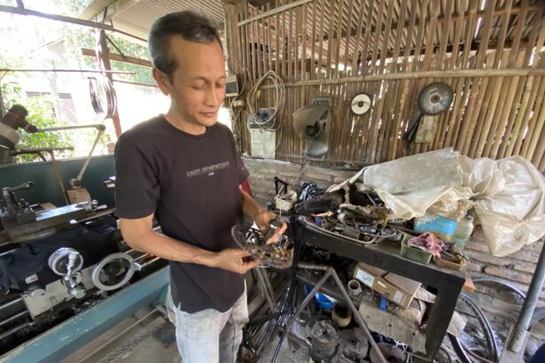 Man wearing a dark T-shirt stands in a cluttered workshop, holding bicycle parts over a workbench with tools and machinery nearby.