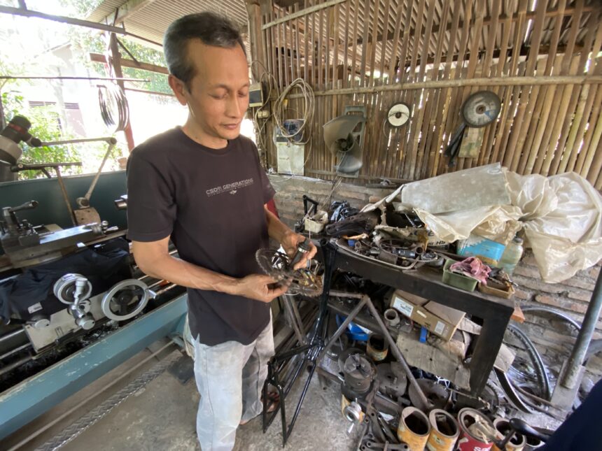 IMG_9842 - Mabur.co Man wearing a dark T-shirt stands in a cluttered workshop, holding bicycle parts over a workbench with tools and machinery nearby.