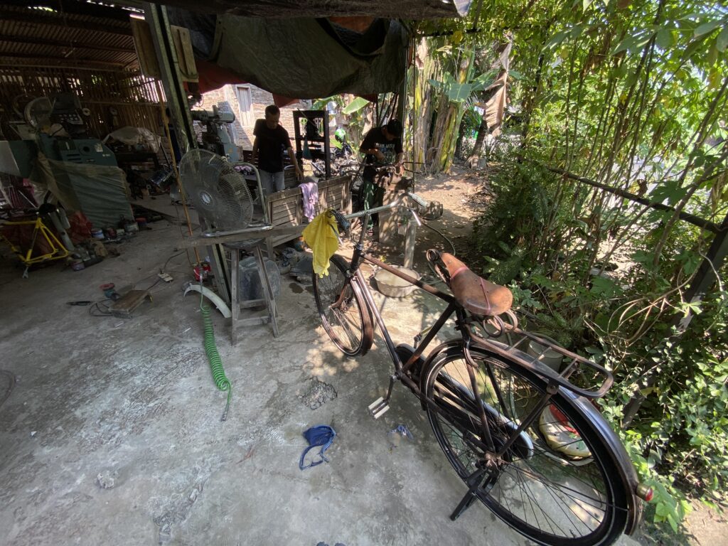 IMG_9850 - Mabur.co Cluttered outdoor workshop with bicycles, tools, and a man at a workbench under a tarp; dense bamboo on the right.
