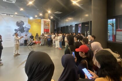 Speaker addressing a crowd in an indoor exhibition; wall graphics show a photographer and text panels behind the group behind him.