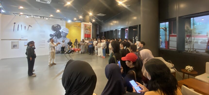 Speaker addressing a crowd in an indoor exhibition; wall graphics show a photographer and text panels behind the group behind him.