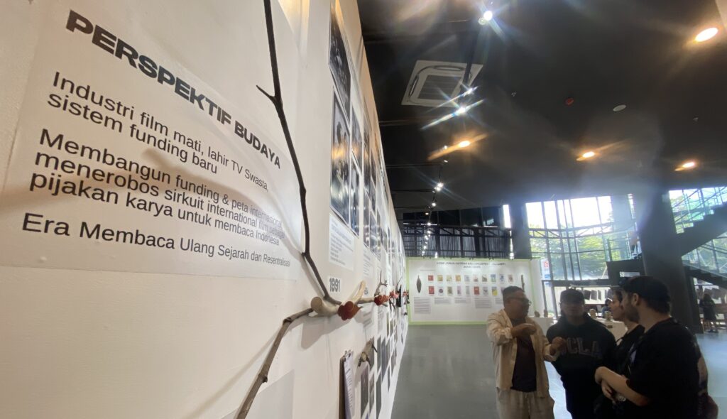 Museum exhibition wall with a large Indonesian text panel and artifacts mounted along the wall; visitors converse in the foreground under gallery lighting.