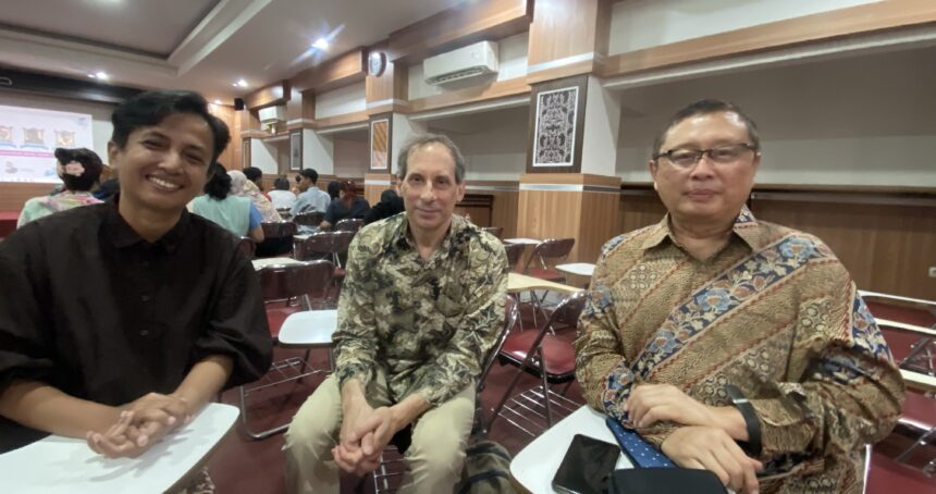 Three men seated in a seminar room pose for a photo; wood-paneled walls and a projection screen visible in the background.