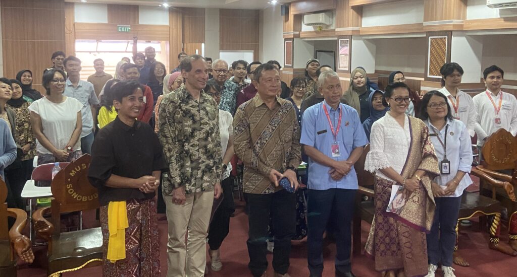 Group of diverse people standing together for a group photo at an indoor event, many in batik and with lanyards in a conference room setting.