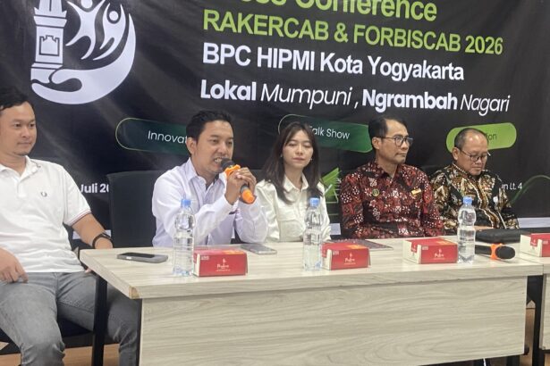 Five panelists seated at a conference table, microphones and water bottles in front, large banner in the background with event text.