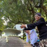 Elderly man in traditional clothing pours water from a jug onto a large ceremonial urn while others assist in a shade-filled outdoor setting.