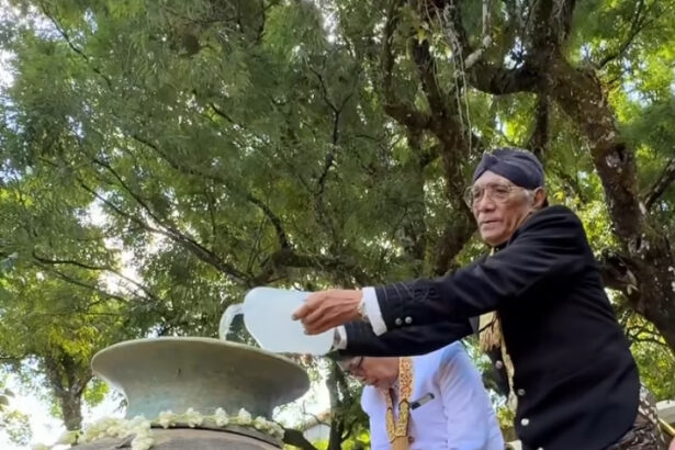 Elderly man in traditional clothing pours water from a jug onto a large ceremonial urn while others assist in a shade-filled outdoor setting.