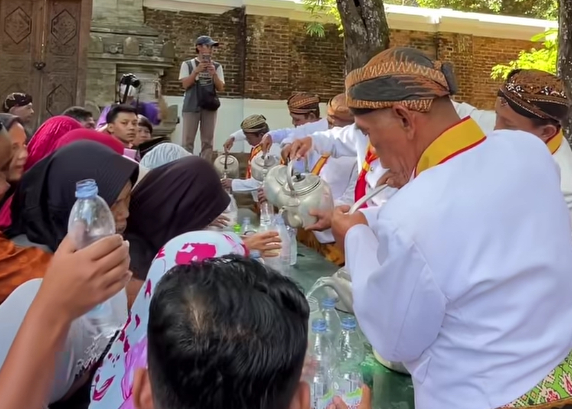 IMG_9989 - Mabur.co Man in white traditional attire pours liquid from a kettle to a crowd at a long table outdoors, with onlookers nearby.