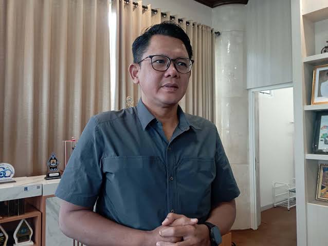 IMG_9993 - Mabur.co Man with short black hair and glasses stands in an office, wearing a blue short-sleeve shirt and a watch on his left wrist.