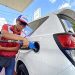 Gas station attendant in red uniform refueling a white van at the pump with a blue nozzle.