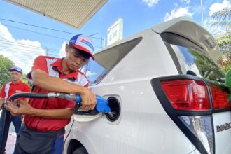 Gas station attendant in red uniform refueling a white van at the pump with a blue nozzle.