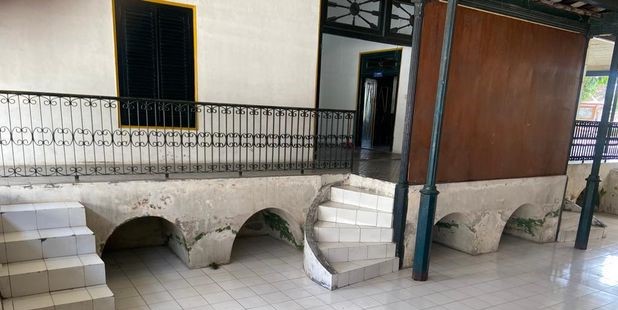 Covered outdoor walkway with arched openings and tiled stairs leading to a doorway, with an iron railing along a low wall.