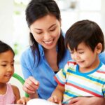 Smiling teacher guides two children as they work on a hands-on activity at a bright classroom table.