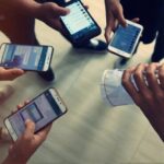 Top-down view of five people holding smartphones around a hand gripping a crumpled white paper bag on a light floor, implying a mobile payment or exchange.