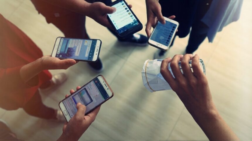 Top-down view of five people holding smartphones around a hand gripping a crumpled white paper bag on a light floor, implying a mobile payment or exchange.