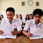 Two male Indonesian students in white shirts with blue ties read papers at a wooden desk, smiling; others sit in rows behind them in a classroom.