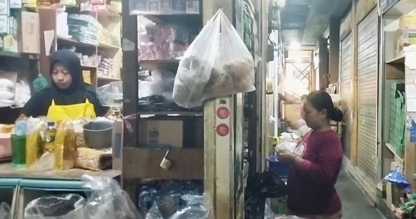 Market stall with a worker in a yellow apron behind a cluttered counter and a customer at a narrow aisle.