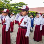 Group of young students in maroon skirts and white shirts, saluting during a school ceremony outdoors.