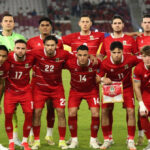 Soccer team in red jerseys posing for a pre-match photo on the field, one player holds a pennant.