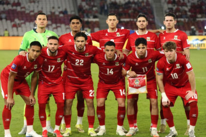 Soccer team in red jerseys posing for a pre-match photo on the field, one player holds a pennant.