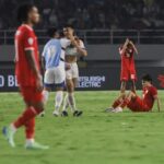 Two red-clad players celebrate with a hug while a teammate sits on the grass with hands on his head, showing disappointment in a soccer match backdrop.
