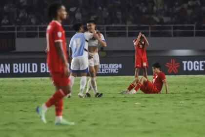 Two red-clad players celebrate with a hug while a teammate sits on the grass with hands on his head, showing disappointment in a soccer match backdrop.