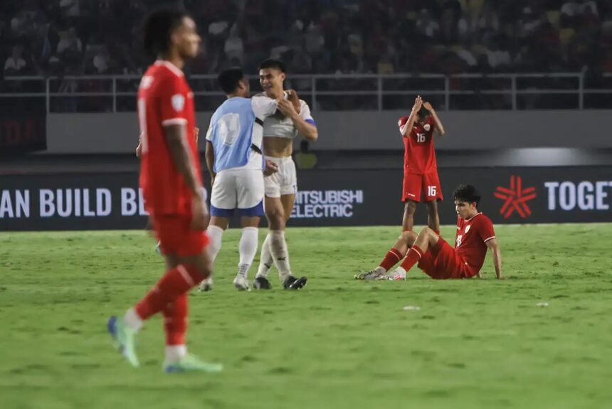 Two red-clad players celebrate with a hug while a teammate sits on the grass with hands on his head, showing disappointment in a soccer match backdrop.