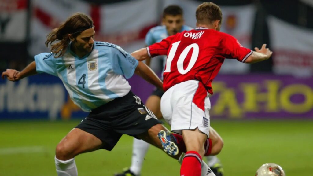 Two soccer players in a duel for the ball on a green pitch: one in light blue Argentina jersey (number 4) and the other in red England jersey (number 10).