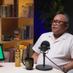 Man in a white polo shirt speaks into a studio microphone during a podcast, seated at a desk with a tablet, bottle, and mug nearby, in a cozy set with bookshelves in the background.