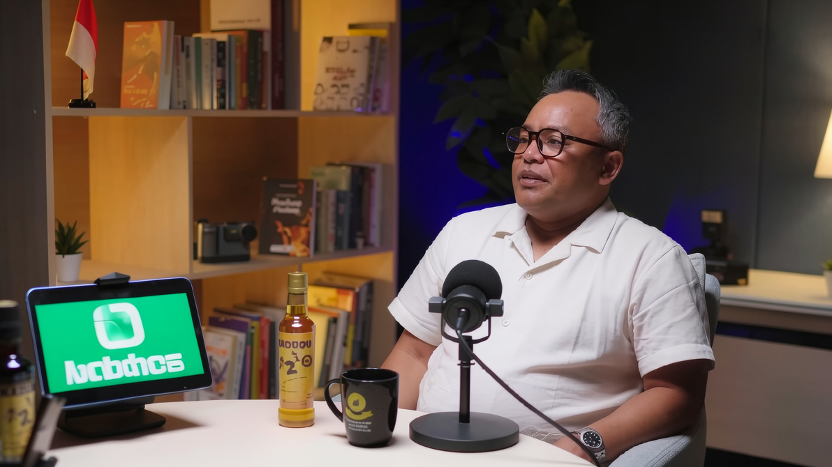 Man in a white polo shirt speaks into a studio microphone during a podcast, seated at a desk with a tablet, bottle, and mug nearby, in a cozy set with bookshelves in the background.