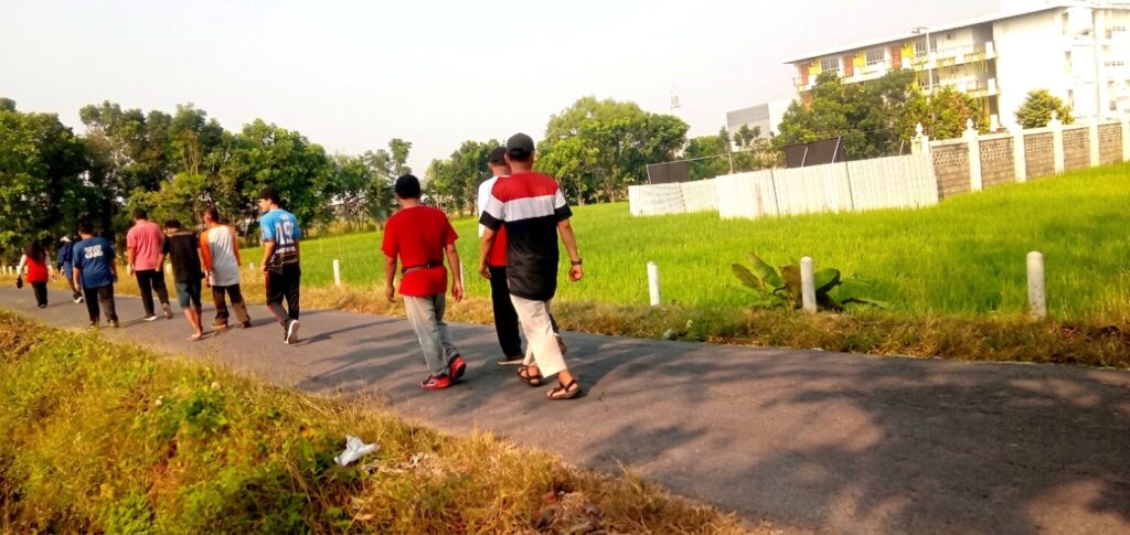 Group of people walking away along a paved path beside green fields, with trees and a white fence in the background and buildings beyond.