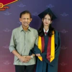 Graduate in teal cap and gown with maroon and gold stole posing with a man in a light striped shirt in front of a school backdrop with a logo banner at the top left.