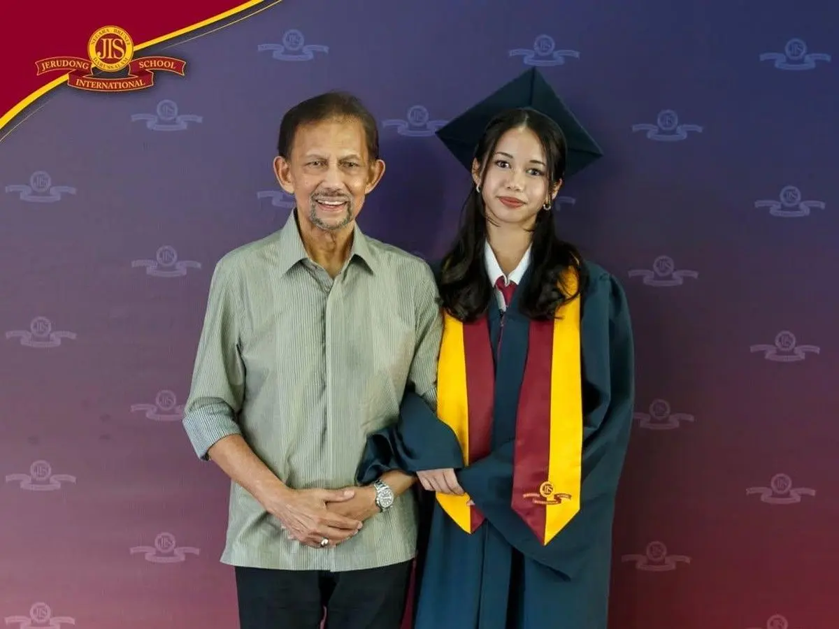 Graduate in teal cap and gown with maroon and gold stole posing with a man in a light striped shirt in front of a school backdrop with a logo banner at the top left.