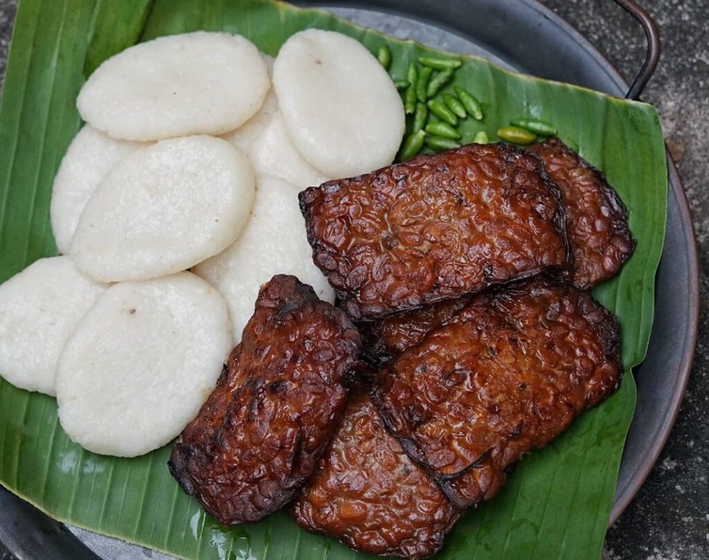 Banana leaf-lined plate with white round steamed cakes and several glazed grilled meat slices, plus a few green chilies in the corner.",