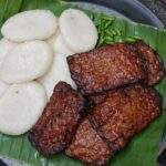 Banana leaf-lined plate with white round steamed cakes and several glazed grilled meat slices, plus a few green chilies in the corner.",