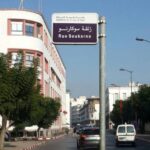 Street scene with a blue and white street sign reading Rue Soukarno in Arabic and French, over a tree-lined urban avenue with white buildings and parked cars.