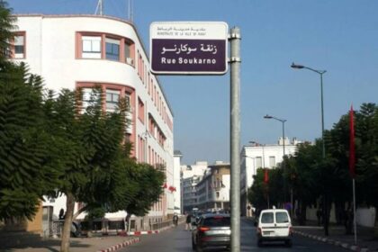 Street scene with a blue and white street sign reading Rue Soukarno in Arabic and French, over a tree-lined urban avenue with white buildings and parked cars.