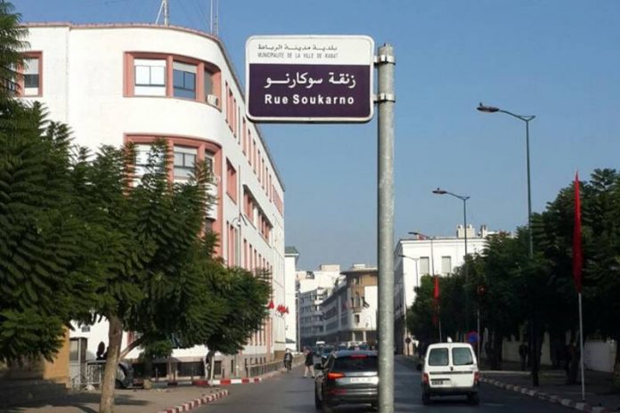 Street scene with a blue and white street sign reading Rue Soukarno in Arabic and French, over a tree-lined urban avenue with white buildings and parked cars.