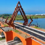 Orange arch bridge with tall rust-colored angular sculpture over a river and a coastal road toward the sea.