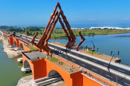 Orange arch bridge with tall rust-colored angular sculpture over a river and a coastal road toward the sea.