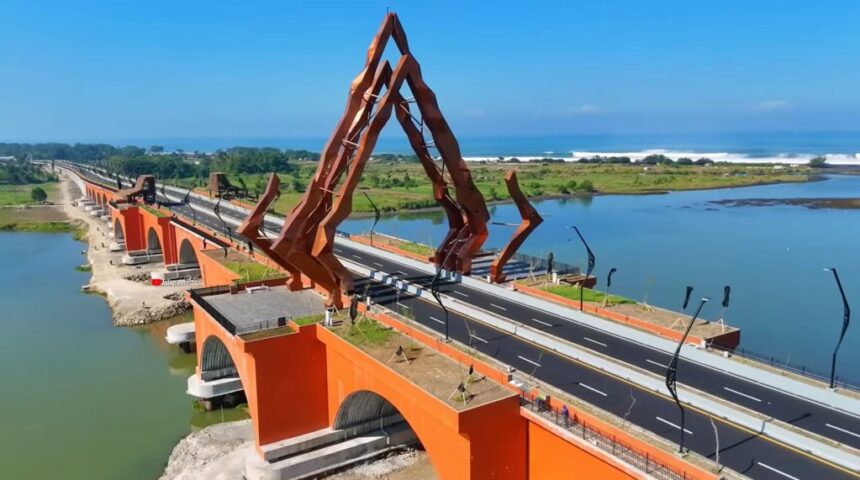 Jalur-Selatan-Jembatan-Pandansimo-Beroperasi-1024x571 - Mabur.co Orange arch bridge with tall rust-colored angular sculpture over a river and a coastal road toward the sea.