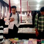 A bookstore or book fair scene with two men browsing books at a long table of stacked titles while a woman in a striped hijab looks through items in the foreground.