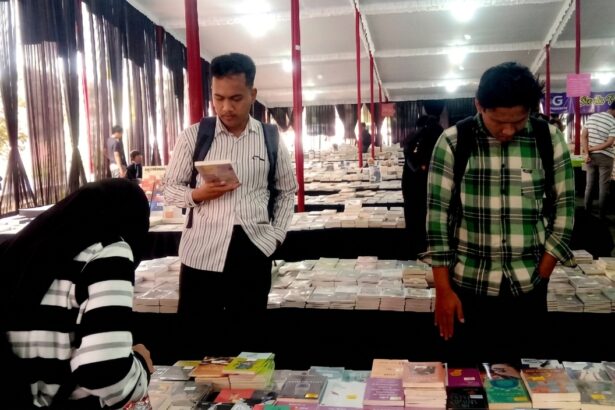 A bookstore or book fair scene with two men browsing books at a long table of stacked titles while a woman in a striped hijab looks through items in the foreground.