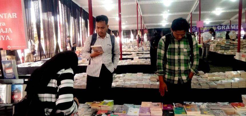 A bookstore or book fair scene with two men browsing books at a long table of stacked titles while a woman in a striped hijab looks through items in the foreground.