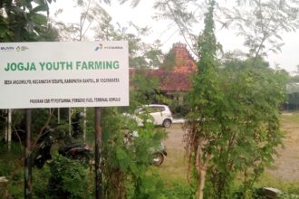 Large outdoor sign reading 'JOGJA YOUTH FARMING' with logos, surrounded by green plants and trees; a building with a red-tiled roof visible in the background