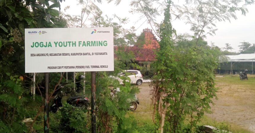 Jogja Youth Farming plang depan bersih FIXX - Mabur.co Large outdoor sign reading 'JOGJA YOUTH FARMING' with logos, surrounded by green plants and trees; a building with a red-tiled roof visible in the background