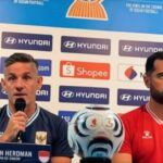 Press conference scene: John Herdman (Head Coach) speaks into a microphone beside a red-shirted teammate, with a soccer ball on the table and sponsor logos in the background.