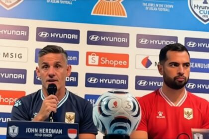 Press conference scene: John Herdman (Head Coach) speaks into a microphone beside a red-shirted teammate, with a soccer ball on the table and sponsor logos in the background.