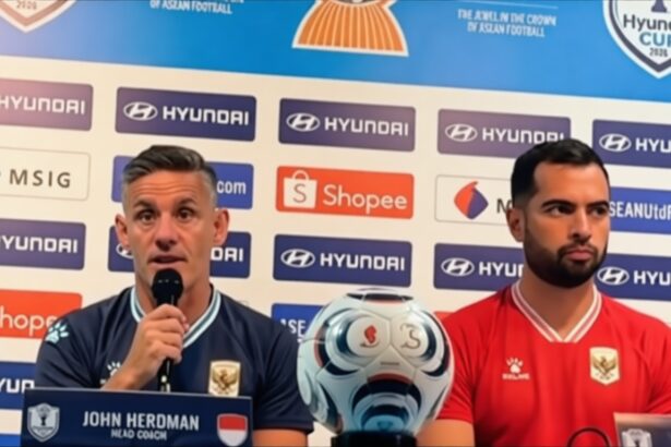 Press conference scene: John Herdman (Head Coach) speaks into a microphone beside a red-shirted teammate, with a soccer ball on the table and sponsor logos in the background.